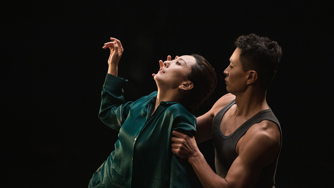 Against a black backdrop, a woman wearing a dark teal shirt leans backwards, one hand raised. Behind her, a man grips her arm while placing his other hand by her face.