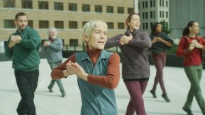 A group of people of various ages stand with one leg in front of the other, twisting their bodies towards the camera. They hold their hands over their hearts, elbows reaching out, and their mouths are open as they sing. They wear a mix of red, purple, green and denim clothing.