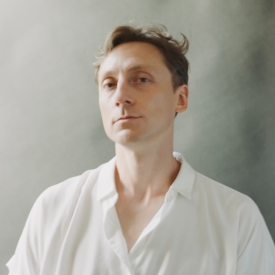 Reid is a Caucasian male with wavy light brown hair and pale skin. He is clean shaven with a neutral expression and wearing a white short sleeve button up shirt with 2 buttons undone. He is sitting in front of a grey paper background.