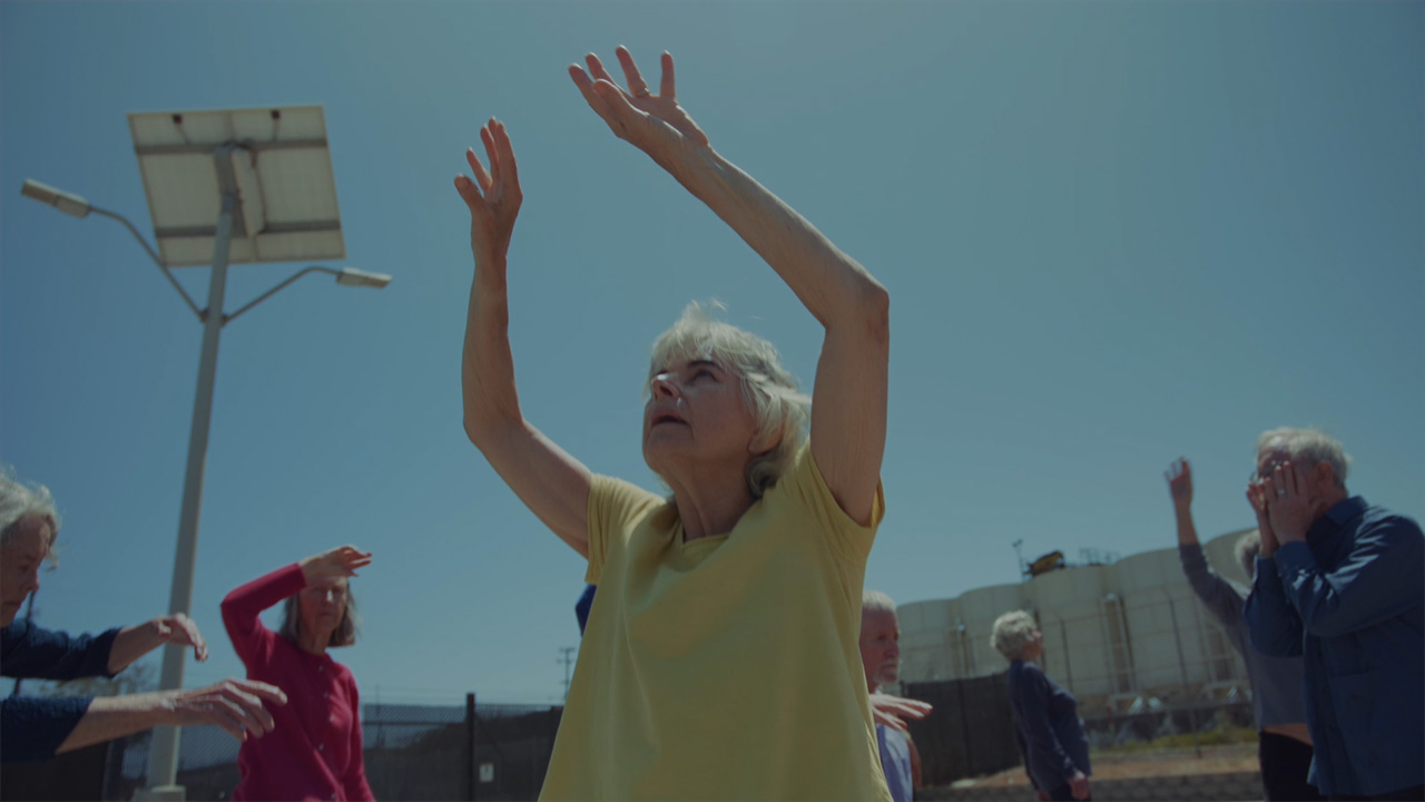 Older adults with Parkinson's Disease dance in an outdoor lot. In the foreground, a woman in a yellow shirt reaches her arms upwards.