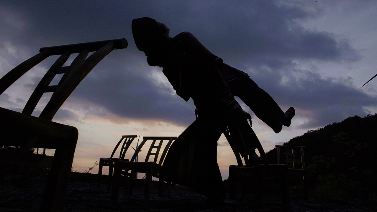 Against a cloudy dusk sky, there are the abstract silhouettes of two dancers leaning back to back. Around them are wooden chairs.