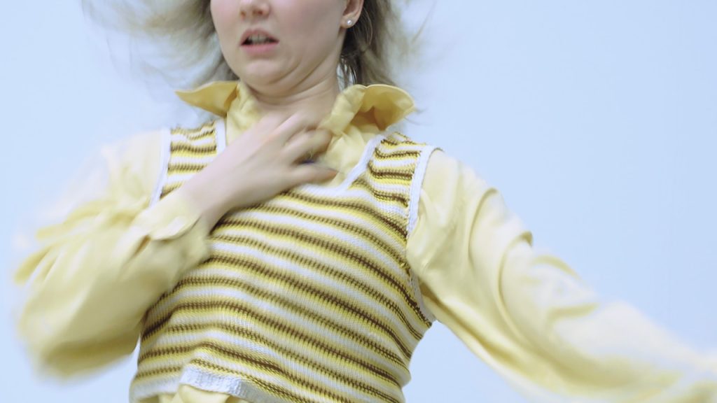 A blonde dancer wearing all yellow moves backwards in a blur, one hand reaching towards their throat.