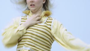 A blonde dancer wearing all yellow moves backwards in a blur, one hand reaching towards their throat.