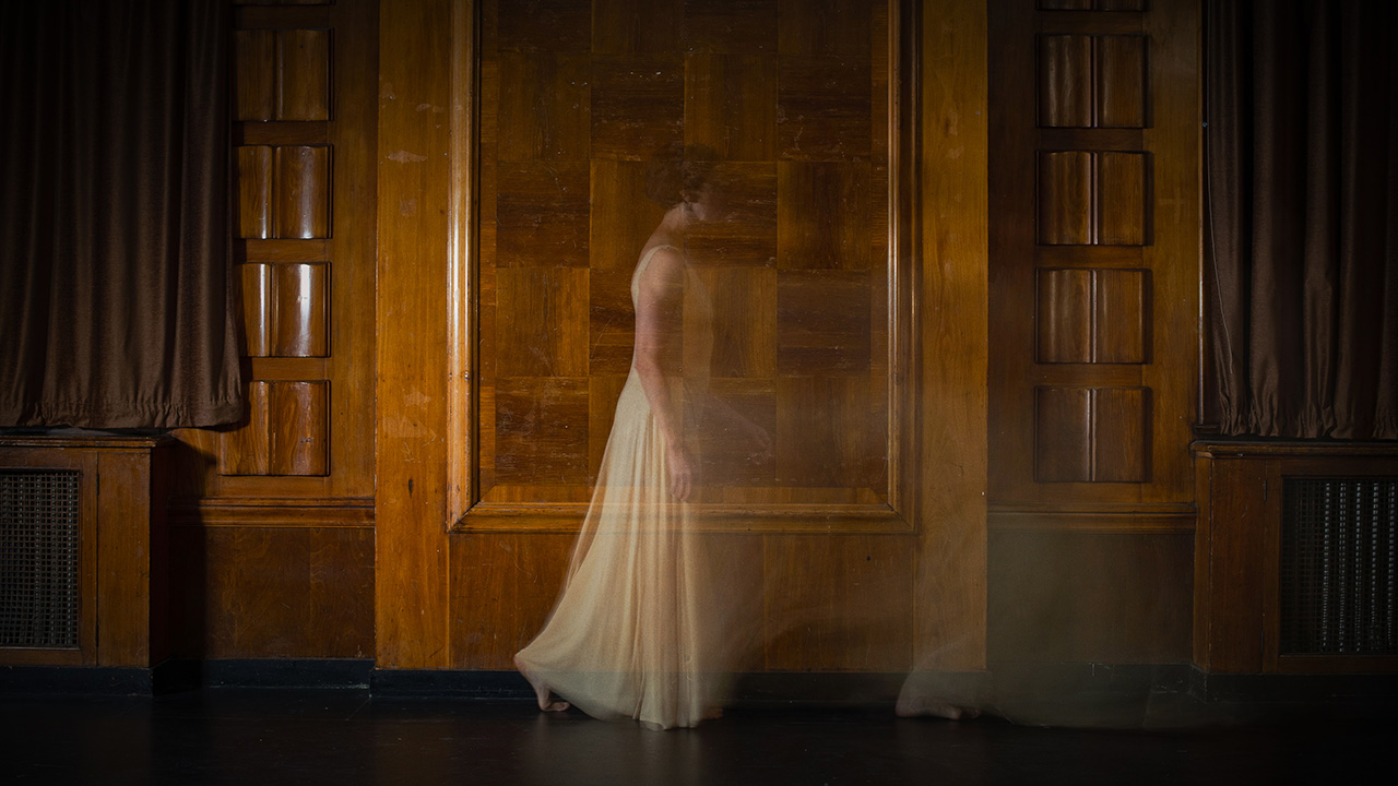 A person in a white dress stands in front of a wooden wall with intricate square patterns in it. They are taking a step forward, and their body is slightly transparent.