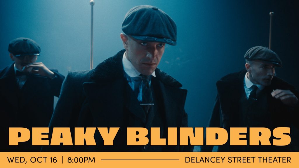 In dim, blue light, a man in a 1910s style hat and suit looks into the distance past the low brim of his hat. His face is intense and angry. Two men stand behind him, looking in the same direction.
