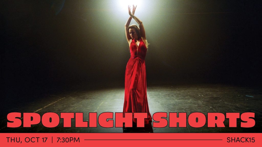 A dancer in a floor-length red dress stands alone on stage, illuminated from the back by a bright spotlight. They pose with their arms above their head, wrists crossed.