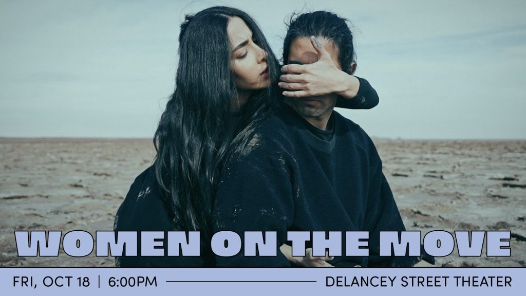 A man and a women, both wearing all black, are on a vast sandy landscape. The woman stands behind the man, covering his eyes with one hand while the other is threaded under one of his armpits.