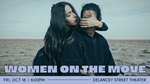 A man and a women, both wearing all black, are on a vast sandy landscape. The woman stands behind the man, covering his eyes with one hand while the other is threaded under one of his armpits.