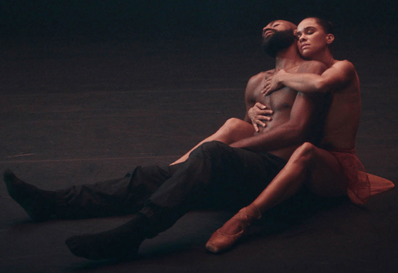 Misty Copeland and Babtunji Johnson sit on a dark floor. Misty sits behind Babatunji, her arms holding his bare chest. He leans his head back on her shoulder.