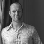 Ben, a white man with short brown hair and a beard, leans against a wall to pose for the camera. He has three silver hoop earrings: one in his right ear and two in his left. He is wearing a white button-up shirt with vertical light blue and dark green stripes. 