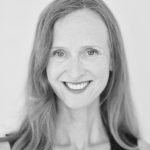 Headshot of Greta, a white woman with long red hair who smiles at the camera. She is wearing a black shirt and stands against a white background.