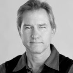 Headshot of Randall Heath, a white man with short blondish hair and blue eyes who smiles at the camera against a simple gray background. He wears a collared button-up shirt and a dark vest.
