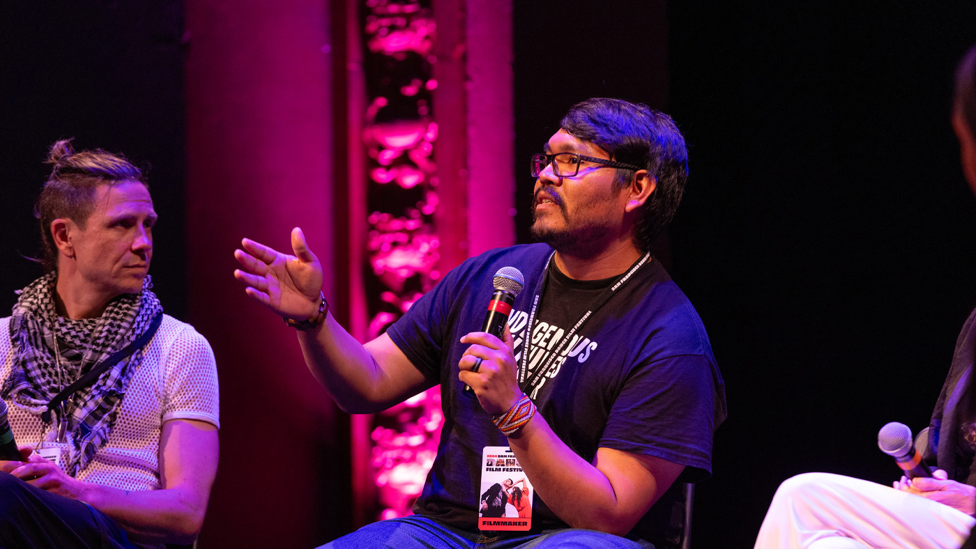 Filmmaker Wade Adakai talking at the Raising Voices Q&A. Filmmaker Wade Adakai is holding a microphone and talking with his hand outstretched in front of him. He wears a shirt that says Indigenous Futures 4EVER and a lanyard that says filmmaker. The lighting on the stage behind him is pink and purple.