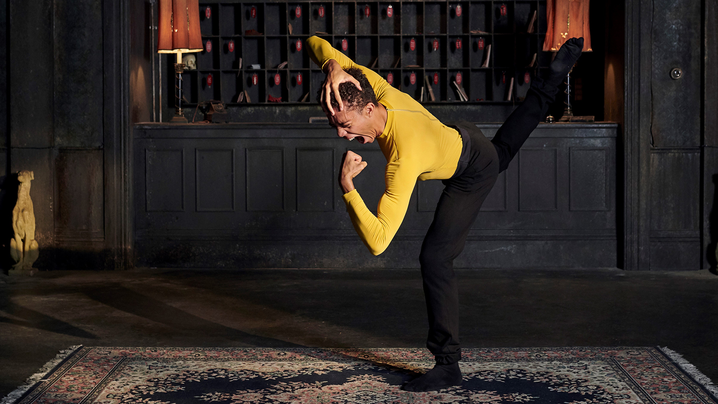 Film still from "I Deeply Know" A dancer in a yellow long sleeved shirt and black pants is in a dark hotel lobby. Behind them is a worn black wall with cubbies, a clock, two mauve old fashioned table lights. The dancer has one hand on their head, and the other is in a fist. They are shouting into their fisted hand, while their back leg is extended up behind them, bent at the knee.