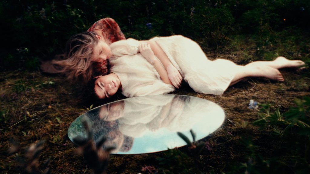 Two people lay on the ground in a forest above a mirror, which reflects their faces and a blue sky. One of them wears a long, white dress, and the other wears red, her hair cascading down one shoulder towards the ground.