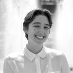 Headshot of Maddy Leitner, a white person with short brown hair, standing in front of two large fountains with a wide smile. They are wearing a white button-down and silver earrings.