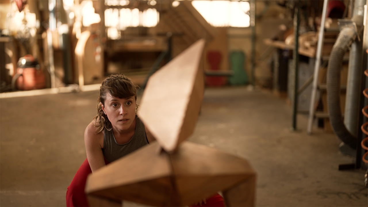 A dancer in a woodwork shop crouches down, only her head, shoulders, chest and part of her legs in sight. Out of focus in the foreground of the image and covering about half of her body from view is a small, unidentifiable wooden structure.