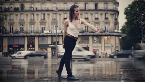 A white woman stands in the rain, with one arm rounded in front of her and her eyebrows furrowed. Cars pass behind her with blurred motion.