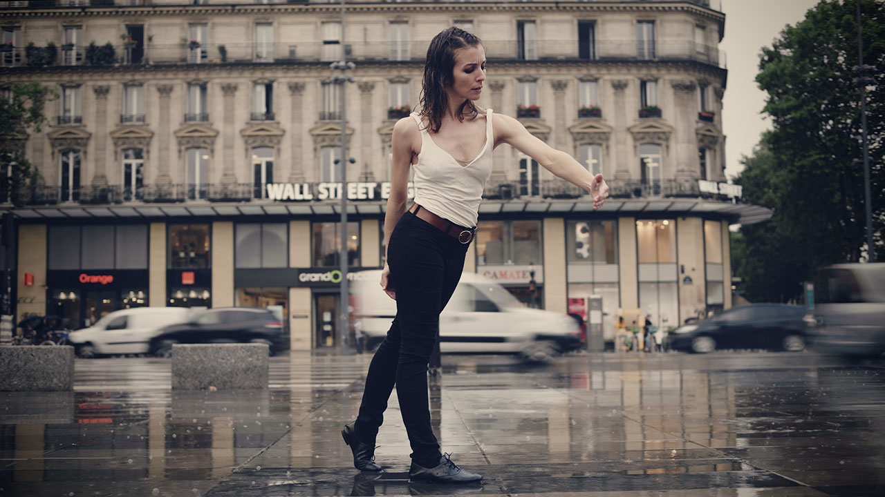 A white woman stands in the rain, with one arm rounded in front of her and her eyebrows furrowed. Cars pass behind her with blurred motion.