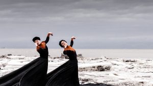 Two dancers in long, black dresses with splashes of orange stand on an ice sheet on the edge of a large body of water. Their left arts extend sideways mimicking the neck and head of a bird, while their right hands are held at their lips.