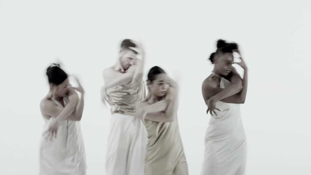 Four dancers in beige and white satin outfits stand in the same pose with their arms twisted around one another, one arm coming up towards their face and the other reaching down towards their waist. Their motion is heavily blurred. The background is flat and completely white. Four dancers in beige and white satin outfits stand in the same pose with their arms twisted around one another, one arm coming up towards their face and the other reaching down towards their waist. Their motion is heavily blurred. The background is flat and completely white.