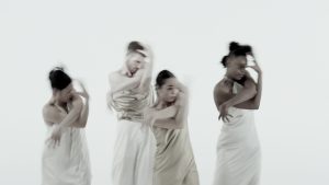 Four dancers in beige and white satin outfits stand in the same pose with their arms twisted around one another, one arm coming up towards their face and the other reaching down towards their waist. Their motion is heavily blurred. The background is flat and completely white. Four dancers in beige and white satin outfits stand in the same pose with their arms twisted around one another, one arm coming up towards their face and the other reaching down towards their waist. Their motion is heavily blurred. The background is flat and completely white.