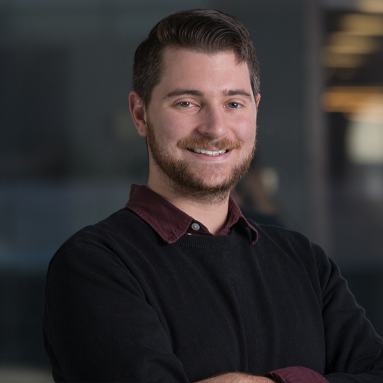Stéphane is a white, European male with dark brown hair and a light brown beard. He is standing in a dark, modern boardroom and wearing a black sweater over a maroon dress shirt.