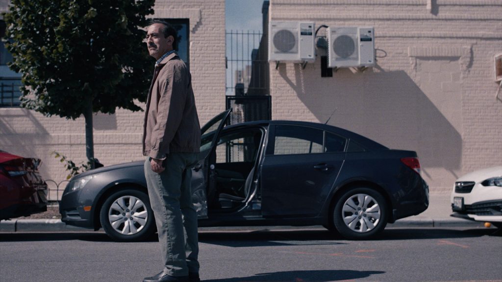 A man stands in a city street with his hands to his sides in front of a car that has one door open. He looks at something off-camera, and his face shows a look of dismay.
