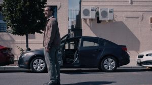 A man stands in a city street with his hands to his sides in front of a car that has one door open. He looks at something off-camera, and his face shows a look of dismay.
