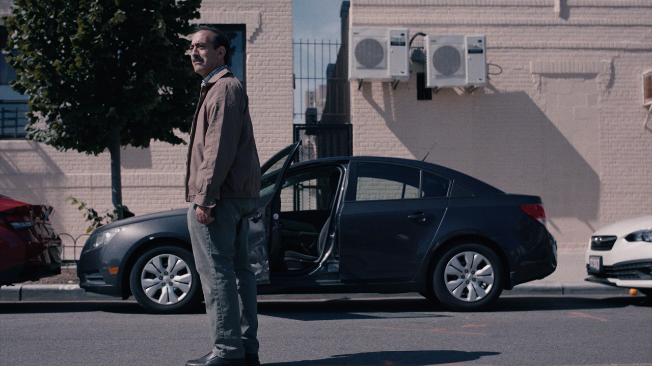 A man stands in a city street with his hands to his sides in front of a car that has one door open. He looks at something off-camera, and his face shows a look of dismay.