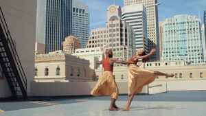 Two dancers in orange tank tops and flowing gold skirts dance on a rooftop in the sun in front of a city skyline. One dancer holds the other's waist, as she leans back with her arms outstretched and one leg extended in attitude behind her.