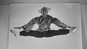 Vintage photo of George Lee, a Chinese male ballet dancer, doing a straddle jump in the air in a photo studio, hands touching his toes. He has a wide smile and wears the original Chinese costume from George Balanchine's Nutcracker: black pants, a flowing top, a woven straw hat, and white ballet shoes.