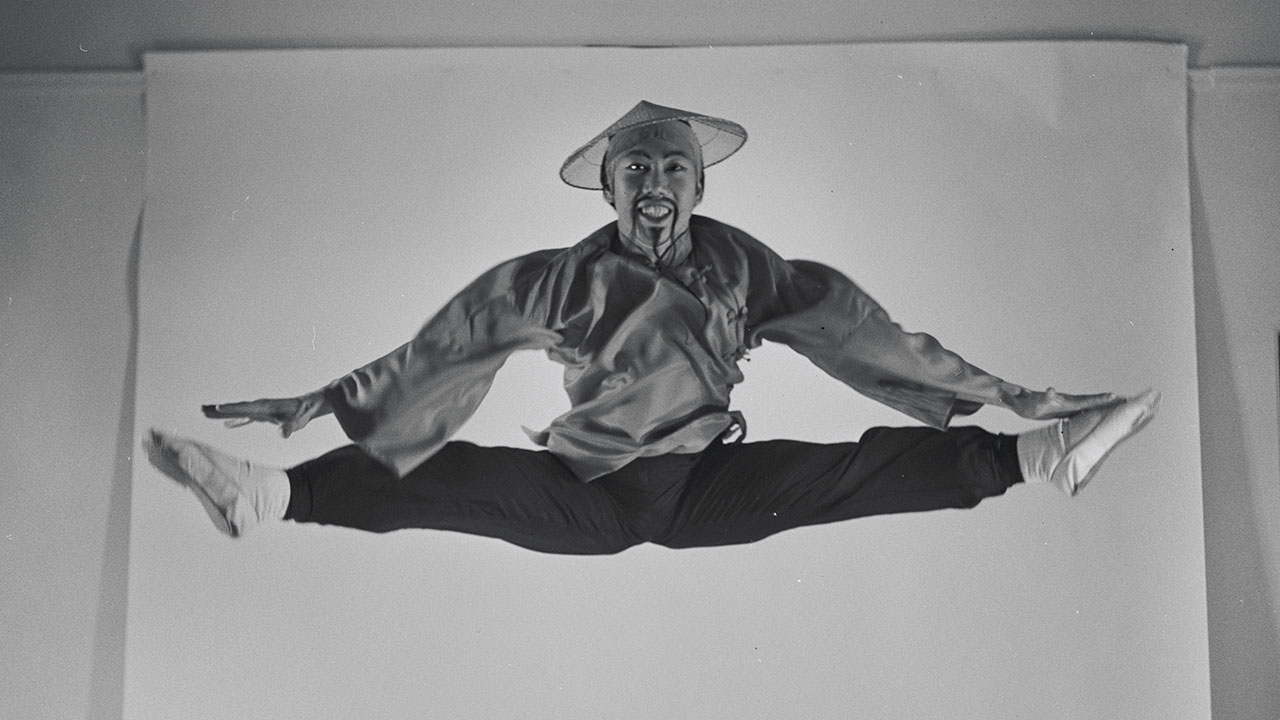 Vintage photo of George Lee, a Chinese male ballet dancer, doing a straddle jump in the air in a photo studio, hands touching his toes. He has a wide smile and wears the original Chinese costume from George Balanchine's Nutcracker: black pants, a flowing top, a woven straw hat, and white ballet shoes.