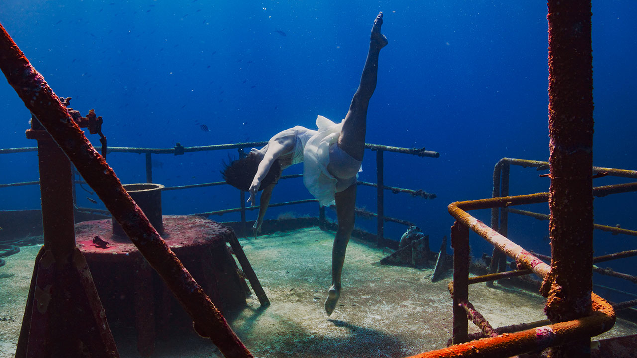 A white woman with flowing brown hair floats underwater above the deck of a wrecked ship covered in algae. She leans backwards in a layout with one leg straight up above her. Fish swim past in the background.