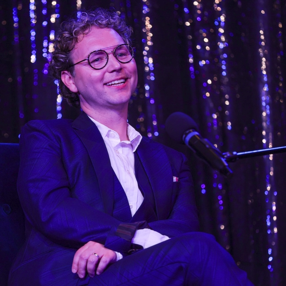Tony has curly blonde hair and black rimmed glasses. He is wearing a navy suit, is smiling, and is seated in front of a sequin curtain with a microphone on a stand in front of him.