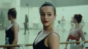 A dancer in bright red lipstick stands at a bar in a ballet classroom. Her eyes are wide and focused, and her reflection in the mirror shows a number pinned to her black leotard. Her hair is slightly frazzled, and a couple flyaways stick out of her bun.