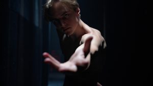 A shirtless dancer reaches an arm towards the camera, his brows furrowed. One light shines on him from above, the rest of the image shrowded in darkness.