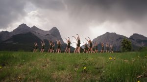 A group of 19 young dancers stand in a grassy field in front of grey mountains and a dramantic sky of grey clouds. They take the same pose: looking over their right shoulder with both arms raised above their heads, hands tilted right.