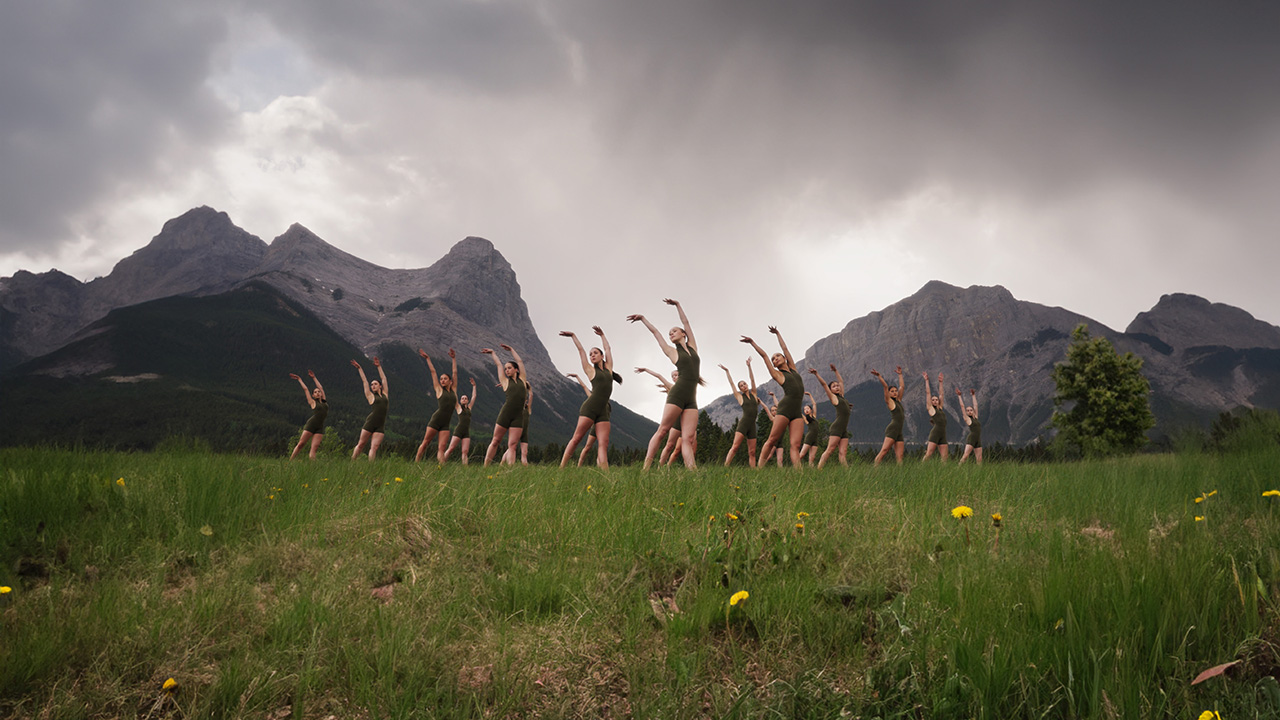 A group of 19 young dancers stand in a grassy field in front of grey mountains and a dramantic sky of grey clouds. They take the same pose: looking over their right shoulder with both arms raised above their heads, hands tilted right.
