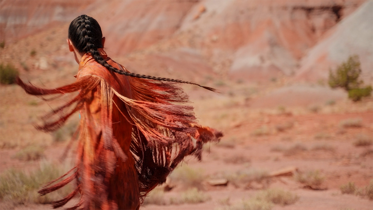 A person with a long braided ponytail is turning away from the camera. They wear an orange jacket with lots of long fringe that hang in the air, the result of movement. They stand in the desert in front of red rocks.