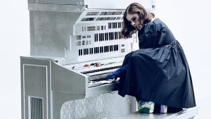 A person in a long blue, puffy dress, blue gloves, and approximately 3-inch platform heels, crouches on a piano bench at a piano that has multiple keyboards extending upwards. They play a chord and look off to the side.