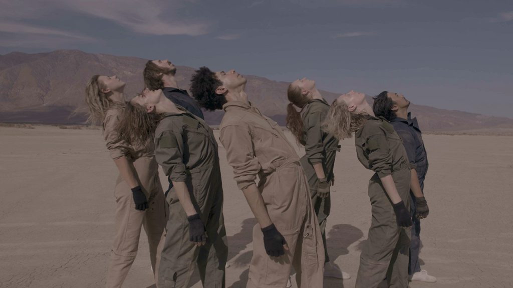 In the desert, a group of seven dancers stand upright and arch back, their faces up toward the sun. They wear an array of dull beige, green and blue jumpsuits and all wear fingerless gloves.