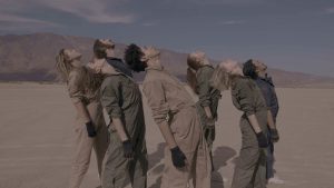 In the desert, a group of seven dancers stand upright and arch back, their faces up toward the sun. They wear an array of dull beige, green and blue jumpsuits and all wear fingerless gloves.