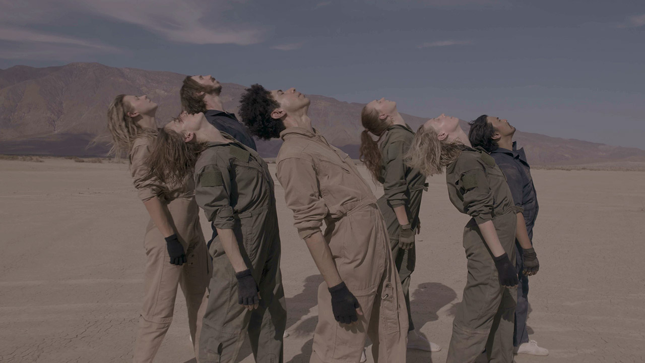 In the desert, a group of seven dancers stand upright and arch back, their faces up toward the sun. They wear an array of dull beige, green and blue jumpsuits and all wear fingerless gloves.