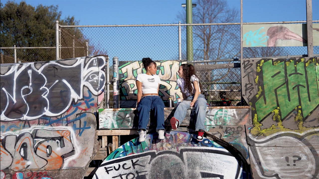 Two dancers sit on a heavily graffitied wall at a skate park, their legs hanging off the edge. They are looking towards each other and laughing.