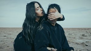 A man and a woman, both with long, black hair and wearing all black, are on a vast sandy landscape. The woman stands behind the man, covering his eyes with one hand while the other is threaded under one of his armpits.