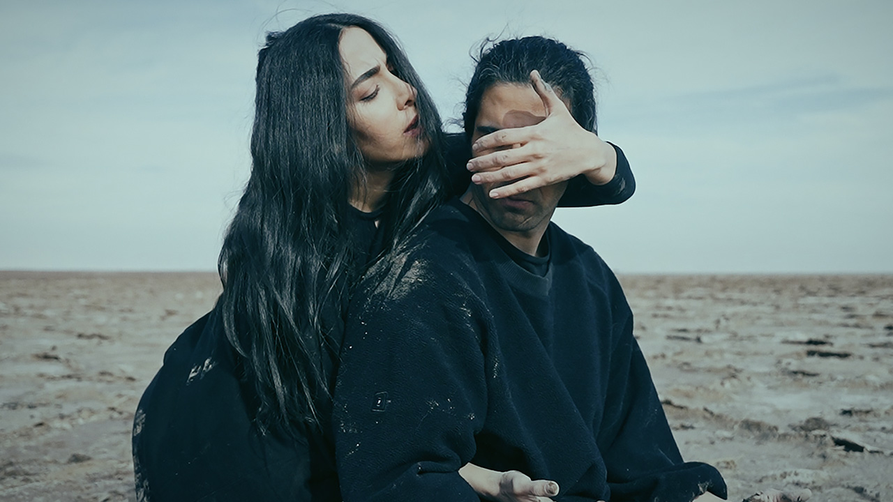 A man and a woman, both with long, black hair and wearing all black, are on a vast sandy landscape. The woman stands behind the man, covering his eyes with one hand while the other is threaded under one of his armpits.