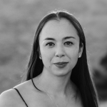 Headshot of Katherine Disenhof, a woman in her mid-30s of Eurasian descent. She has long dark hair and stands in front of a blurry grassy landscape. 