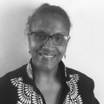 Headshot of Laura Elaine Ellis, a black woman wearing black rimmed glasses. She wears a black shirt that has a geometric pattern around the collar and smiles in front of a white wall.